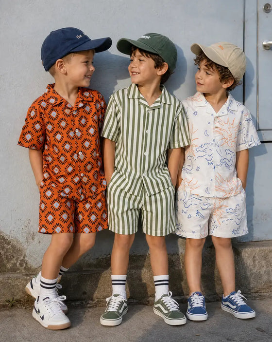 Three boys in matching printed Mumzers shirt-and-shorts sets and caps against a wall