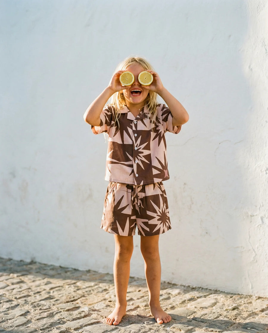 Girl holding lemon slices over her eyes in a brown and pink palm-print Mumzers set against a white wall