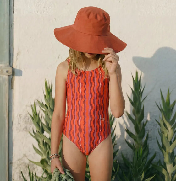 Girl in an orange bucket hat and red wavy-stripe Swimzers swimsuit against a white wall
